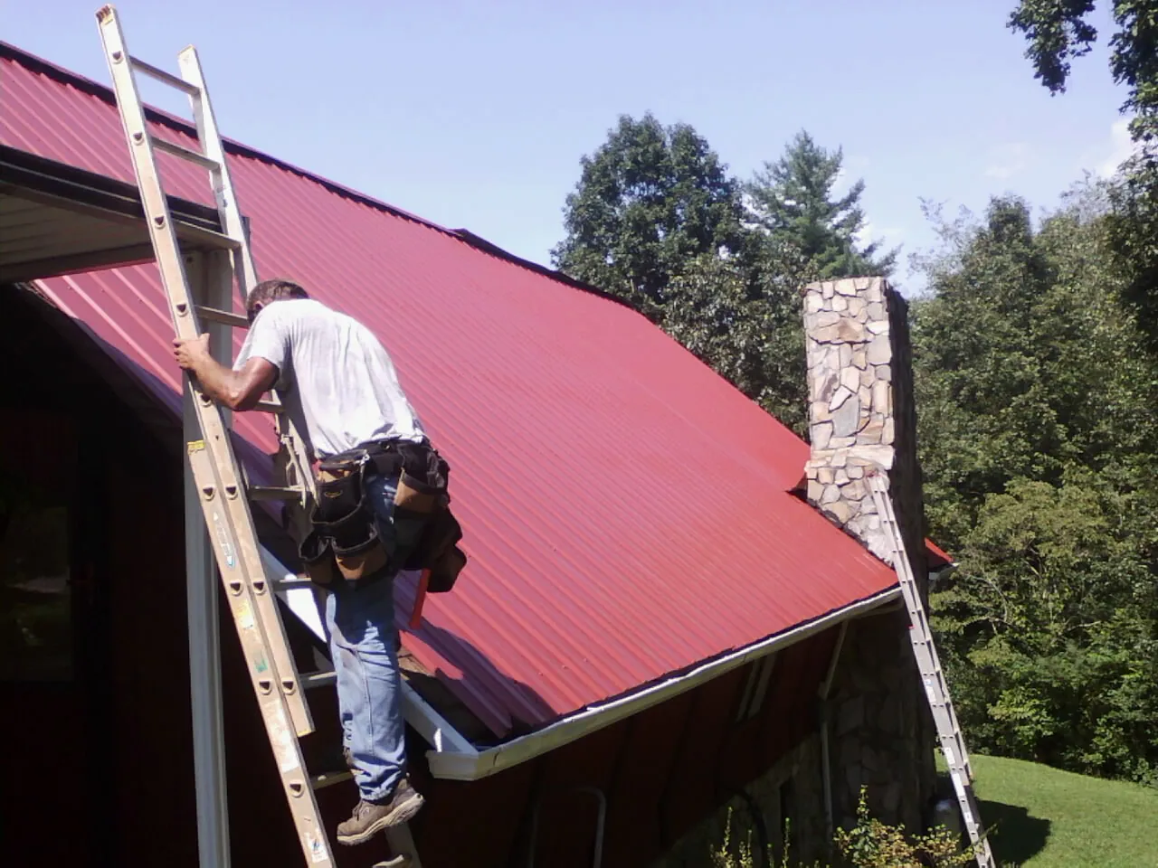 Roofer on a ladder beside a red metal roof and stone chimney — Soffit & Fascia Repair by Roofing Brothers in West Finley